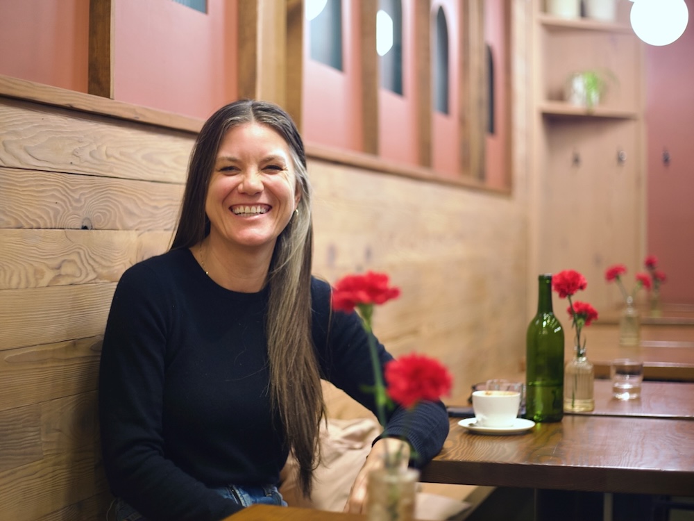 A woman with long salt-and-pepper hair and light skin smiles while sitting at a table in a caf&eacute;. She wears a black sweater and jeans.