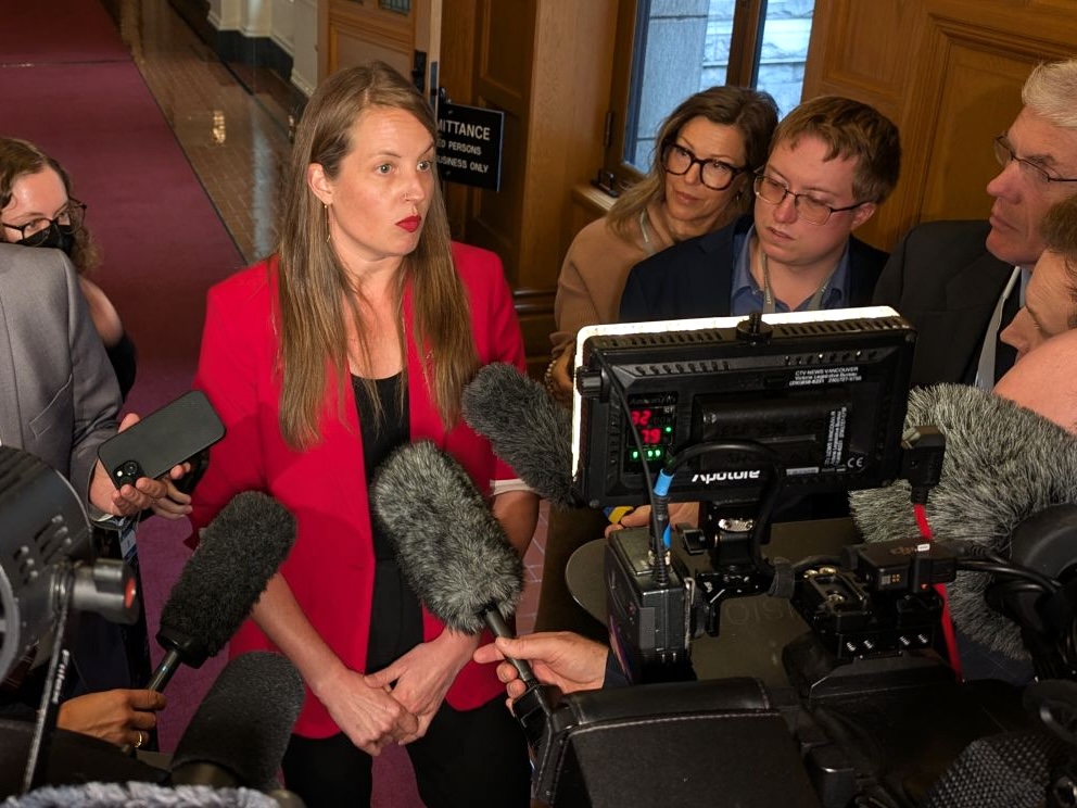 A light-skinned woman in her early 40s wearing a red jacket faces reporters and cameras in a legislature hall.