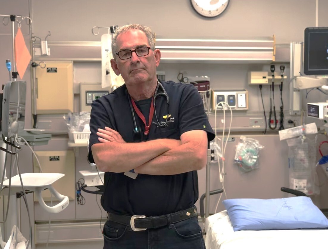 A man in dark blue scrubs with a stethoscope around his neck stands looking at the camera with his arms folded. He has a no-nonsense expression on his face and is standing next to an empty patient bed in a hospital. Various medical devices are hanging from the pale grey walls.