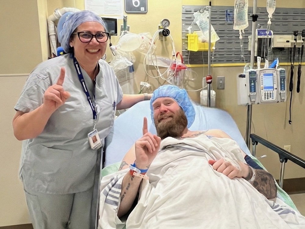 A man with a beard and light skin tone reclines in a hospital bed. Standing beside is a woman in blue scrubs, who will help oversee the surgery. They are both raising one index finger.