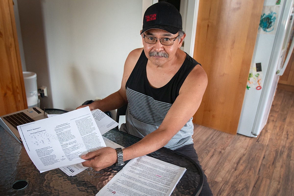 A man with glasses and a moustache, wearing a ball cap that says &lsquo;Tahltan Nation,&rsquo; sits at a table with papers spread in front of him.