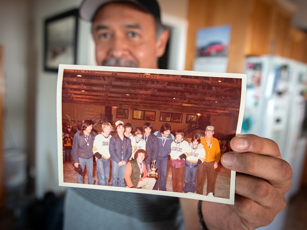 A middle-aged man with a ball cap and moustache holds up a dated photo showing a group of boys and several men posing with a trophy.