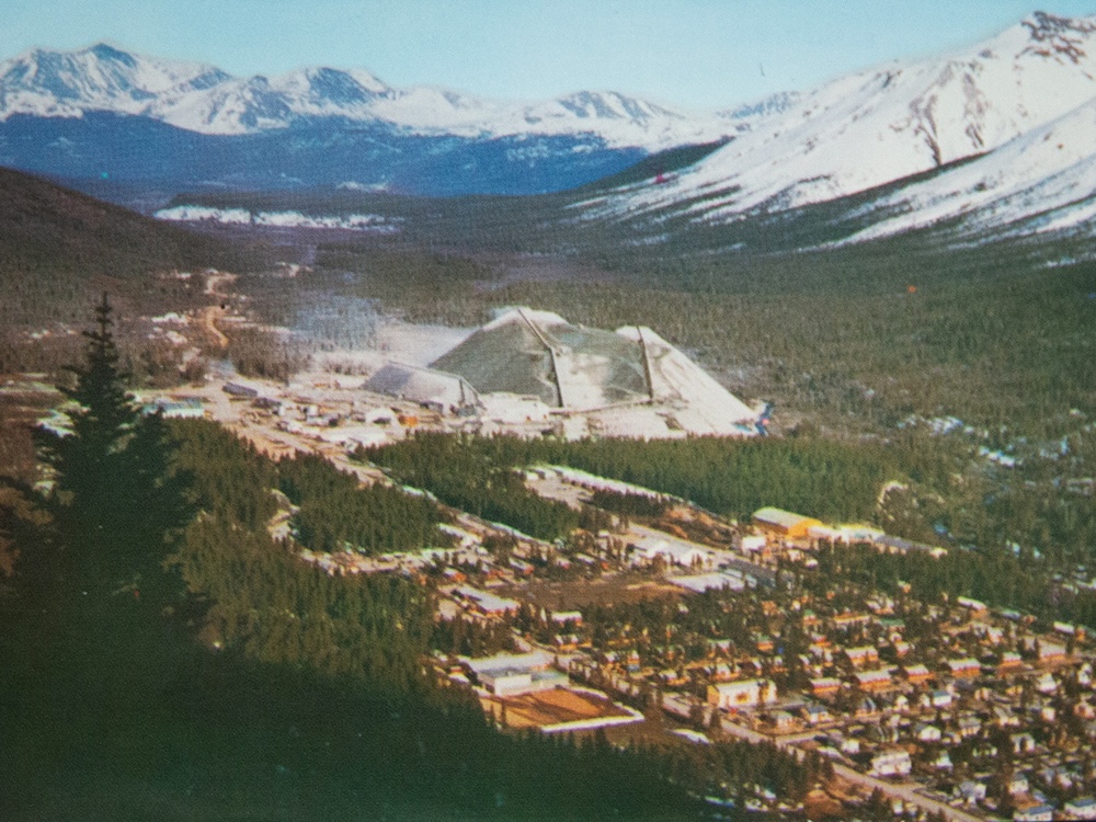 An aerial view of a small community nestled into a mountain landscape. Beyond the community is a large, grey pile of mine tailings that dwarfs the town.