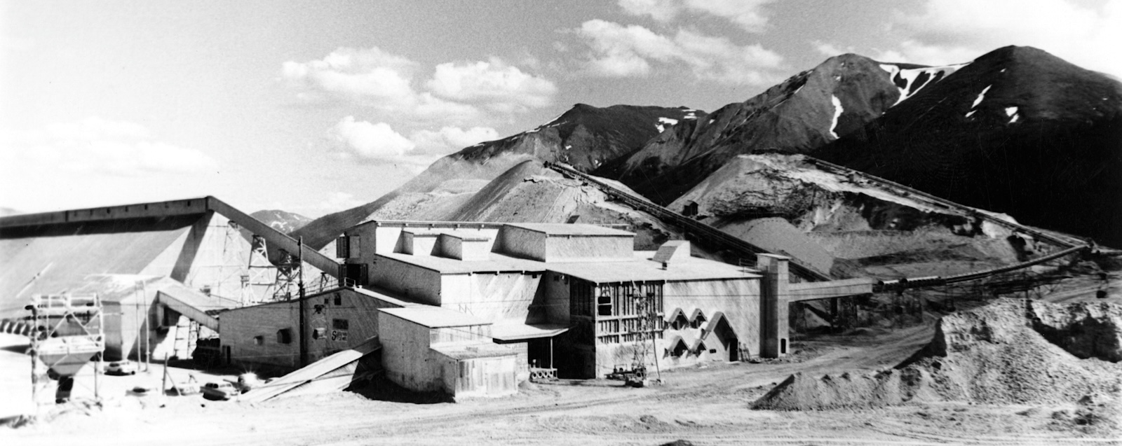 A building for dry rock storage is shown on the left, a mill building is in the foreground, and tailings piles can be seen behind the mill.