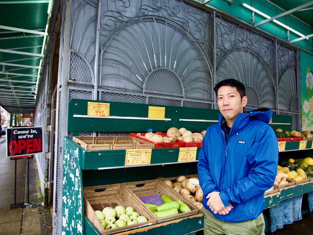 A man with short black hair stands with clasped hands in front of vegetables displayed for sale outside a grocery store.