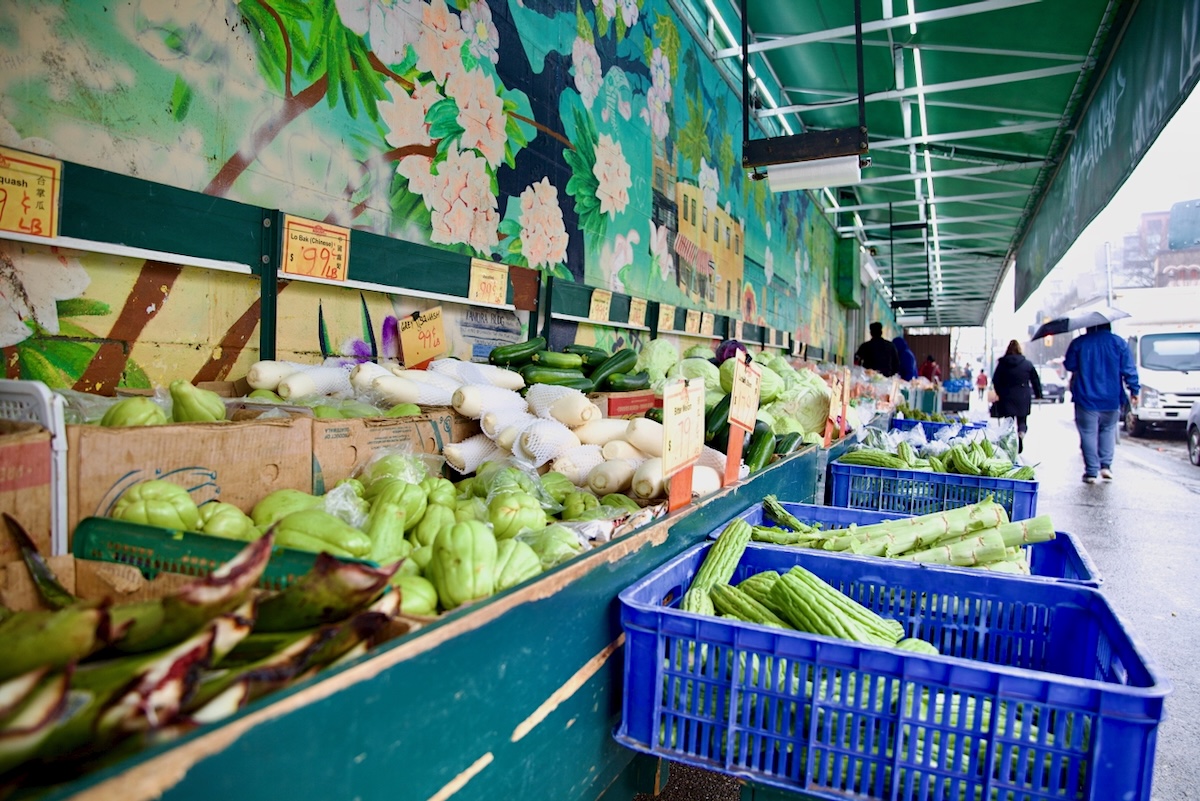 Vegetables are displayed in outdoor shelves and bins outside a grocery store in the Downtown Eastside. A mural of blossoms can be seen on a cinder block wall.