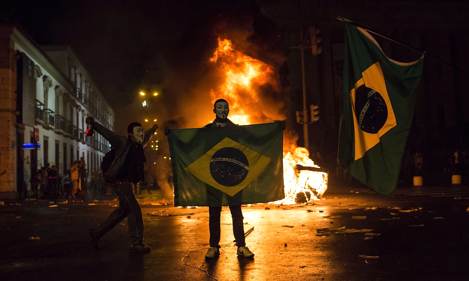 Two protesters wearing Guy Fawkes masks, one holding a Brazilian flag, stand in the street in front of a bonfire in the middle of a street at night.