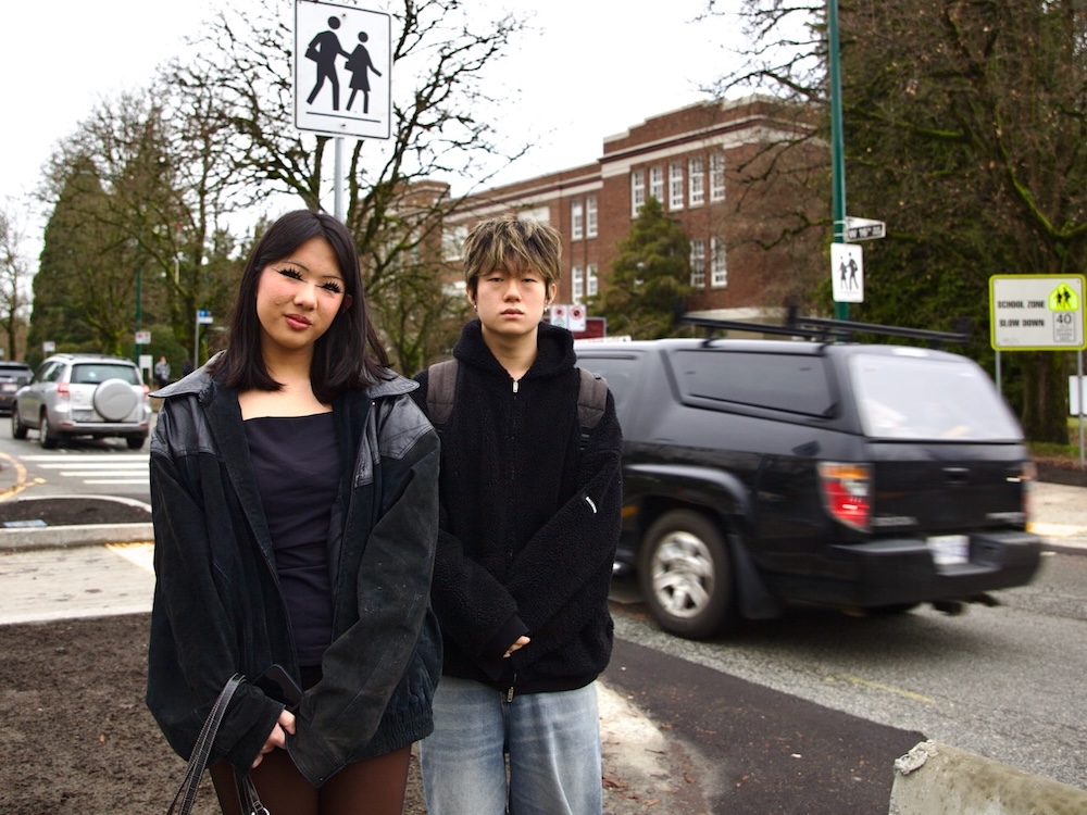 A teenage girl with long black hair and bangs, wearing a black coat and black shirt, and a teenage boy with shaggy blond hair, wearing loose jeans a black hoodie and backpack stand together in front of a busy road in Vancouver. A black car can be seen passing behind the two teens, and in the background is a brick school building.