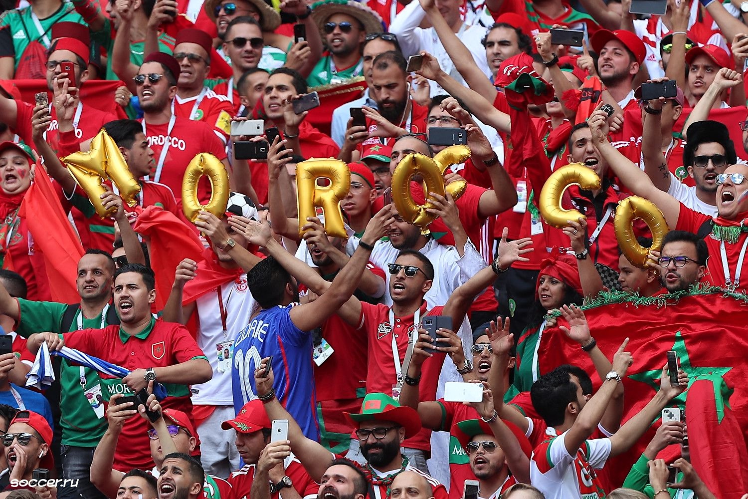 Wildly cheering fans in red jerseys hold up gold letter balloons spelling out &lsquo;Morocco.&rsquo;