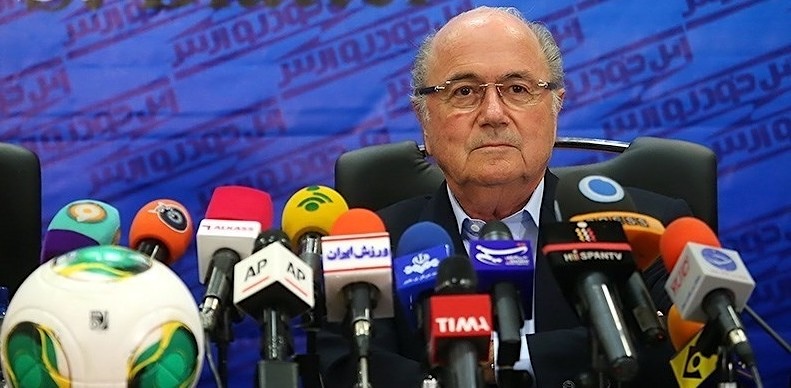 A light-skinned, balding man in a suit sits in front of many microphones.