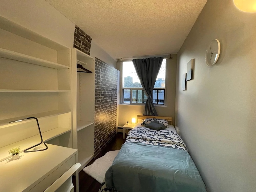 A single bed in a room with a window at one end. One wall of the room is made up of bookshelves.