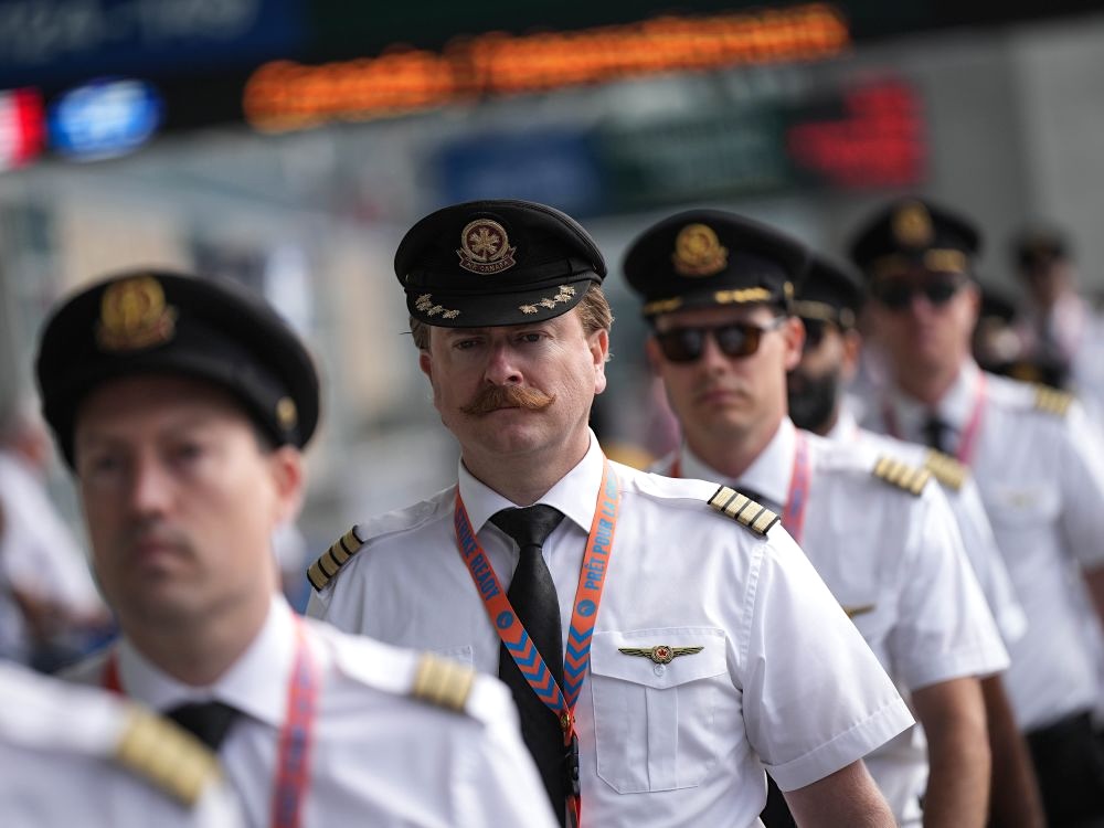 Seven male airline pilots walk in a line. They wear their airline uniforms; one man has a handlebar moustache while another wears aviator sunglasses.