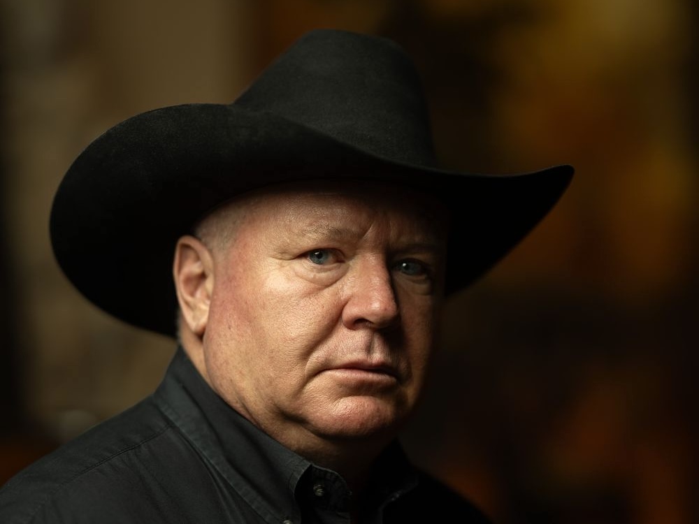 A white, late-middle-aged man with a round face and piercing eyes wears a black cowboy hat.