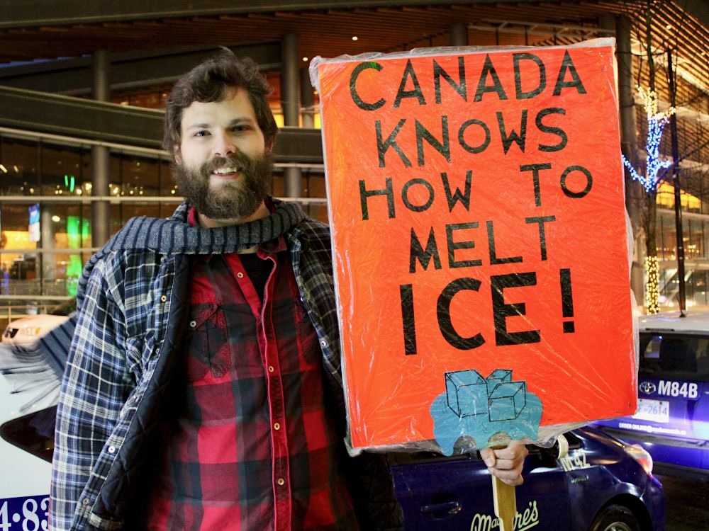 A man with brown hair and a full beard wearing a plaid jacket and scarf holds an orange sign that reads, &lsquo;Canada knows how to melt ICE!&rsquo;