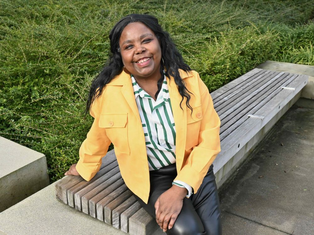 A woman with a yellow jacket and green and white striped shirt smiles at the camera. She has bronze skin and black wavy hair and is sitting in front of a green bush.