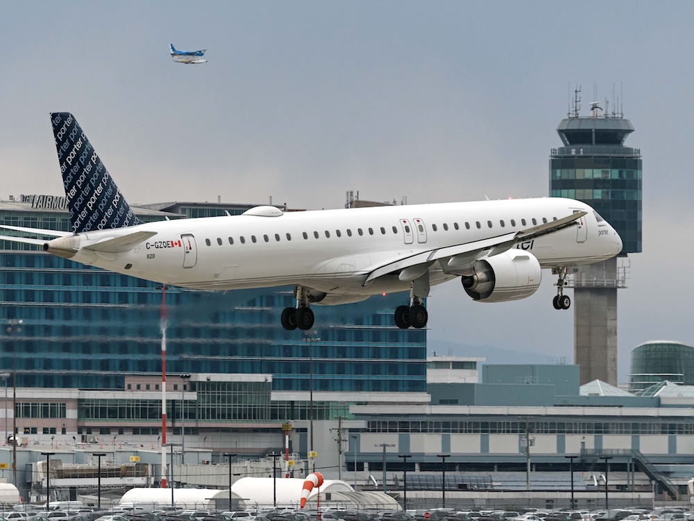 A white passenger plane hovers over the ground with an air traffic control tower behind it.