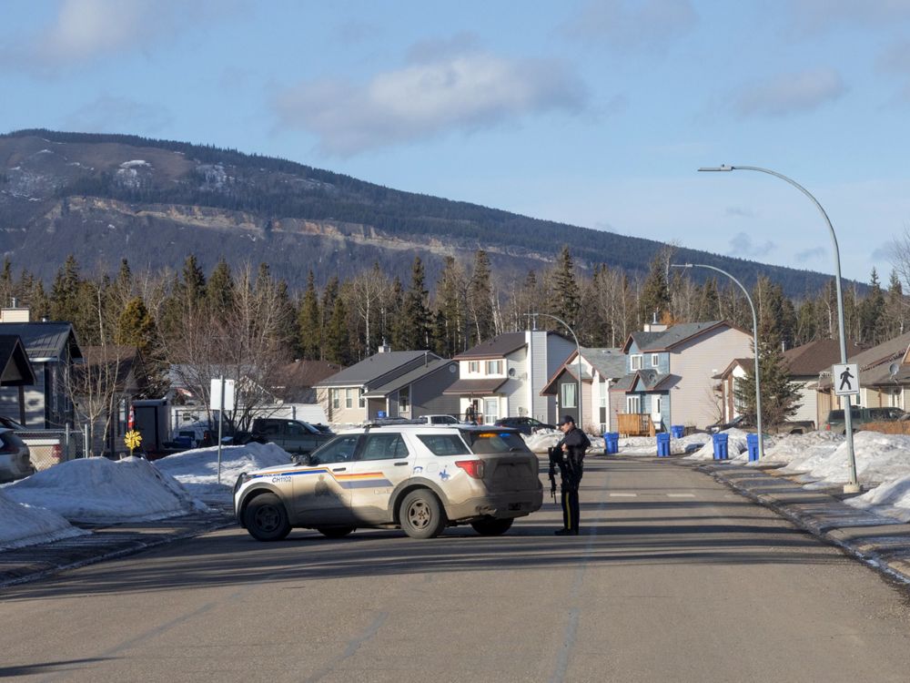 An RCMP officer armed with a rifle is parked diagonally across a suburban-looking street. There is a low mountain in the background and snow on the ground. 