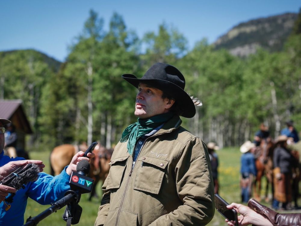 A man in a black cowboy hat wearing a green scarf speaks to reporters in front of mountains.