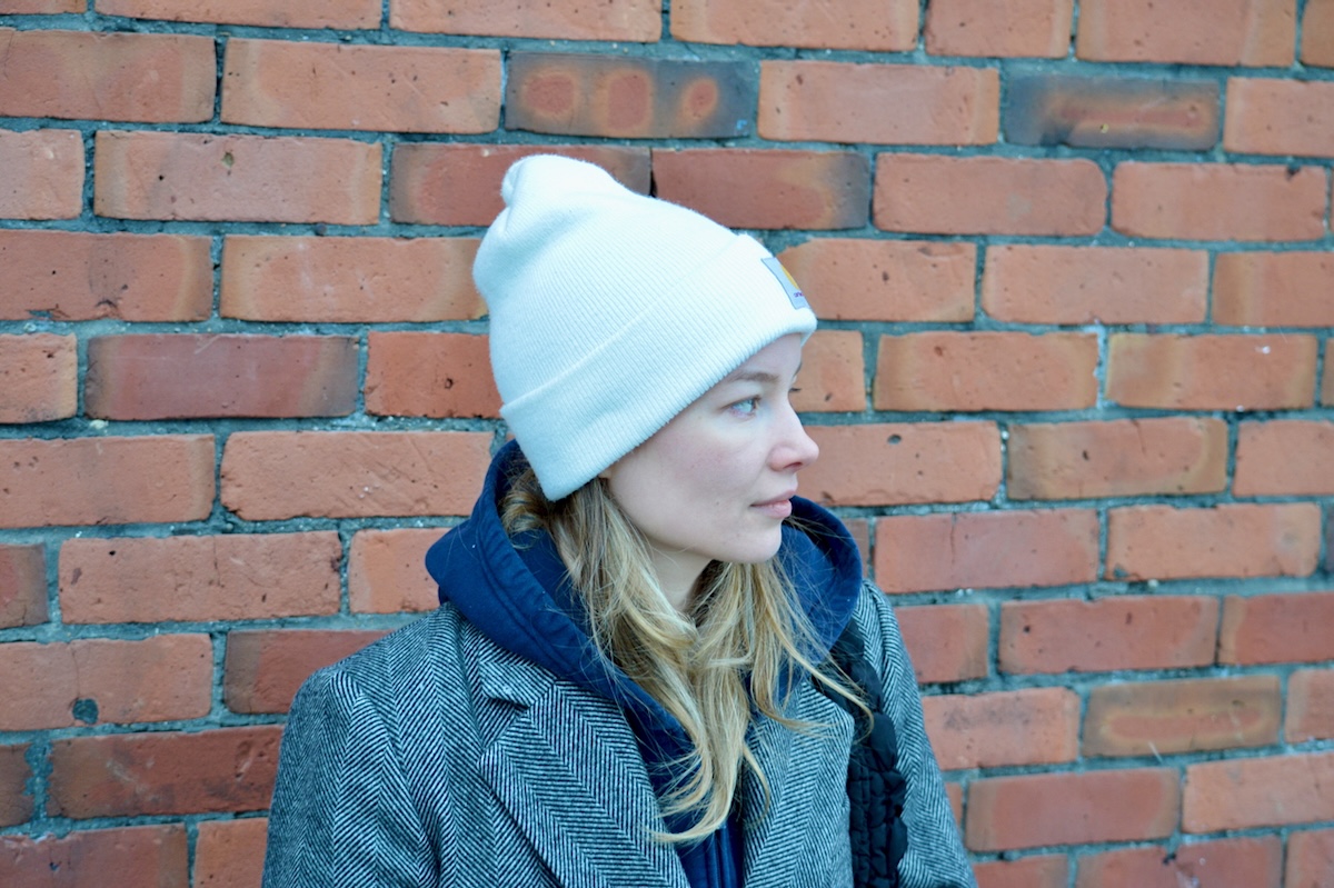 A woman in her 20s with blond hair and a wearing a white toque sits against a brick wall, looking to the right.