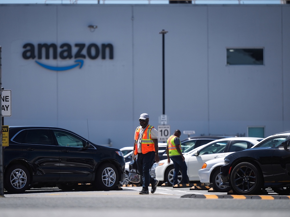 A parking lot with a large boxy building in the background. The building is white and has an Amazon logo. In the foreground, a worker wearing an orange safety vest can be seen walking through the parking lot.