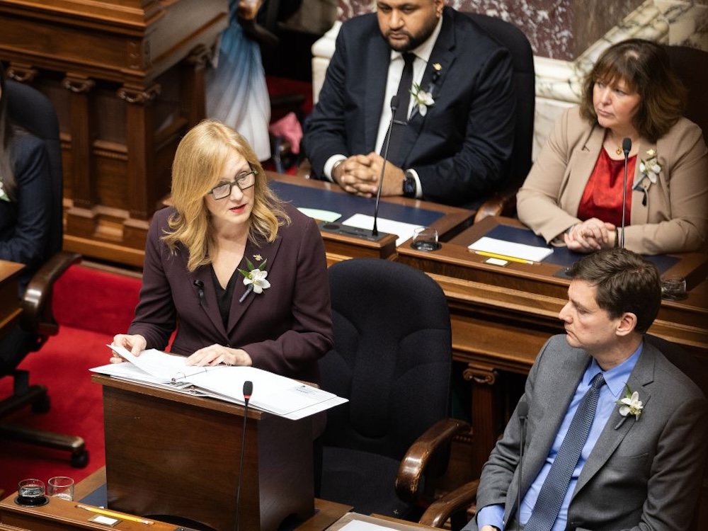 A photo shows Brenda Bailey, in a burgundy suit, standing and speaking as David Eby and other MLAs look on.