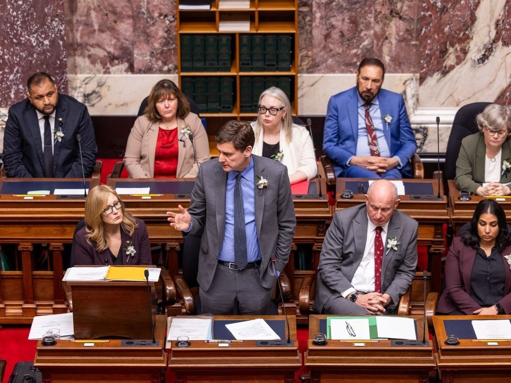 A tall light skinned middle-aged man with a grey suit and brown hair stands and gestures among seated people with a marble wall behind.