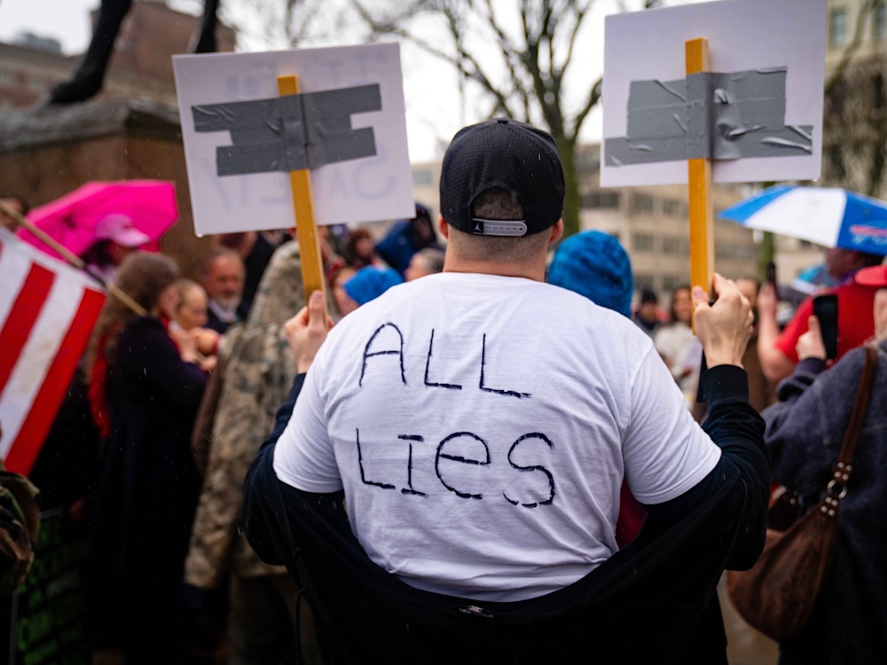 A person at a protest, viewed from behind, holds two signs, wearing a white T-shirt with &lsquo;All Lies&rsquo; written on the back.