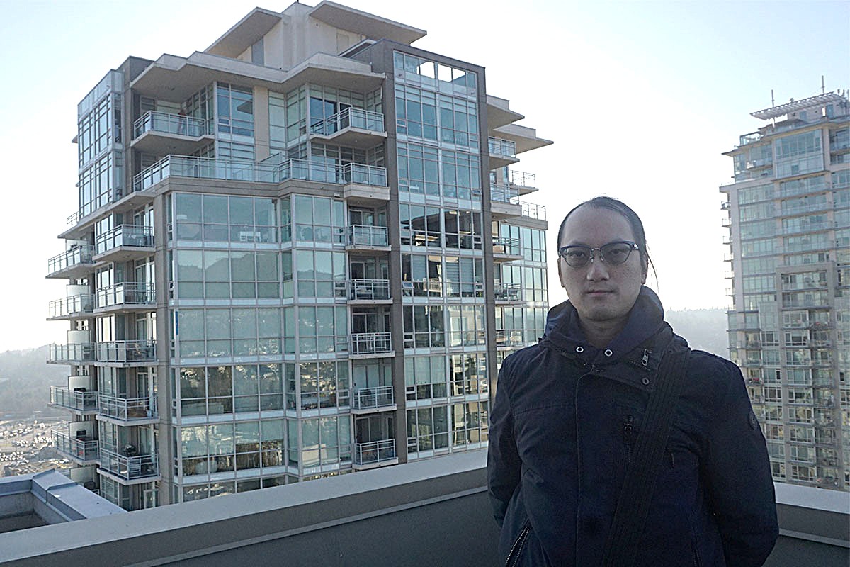 Simon Ng has long hair and is standing on top of a tall building with other condo buildings behind him.