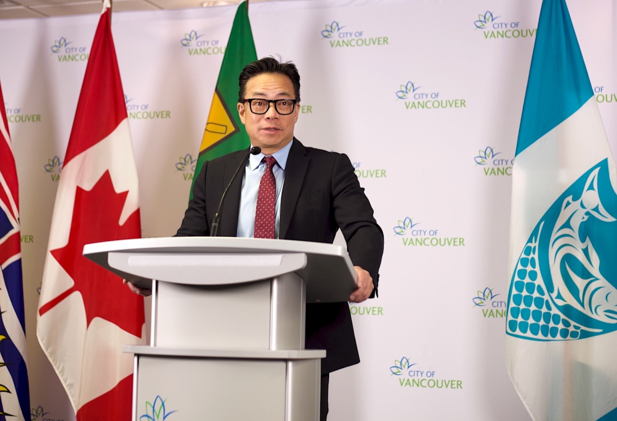 A middle-aged man with short black hair stands at a lectern with various city and provincial flags in the background.