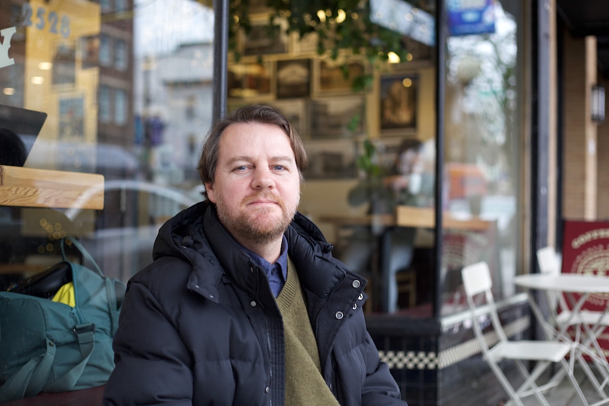 A middle-aged man with brown hair and a beard and moustache sits at a table outside a caf&eacute;. He wears a puffy black coat and a green sweater.