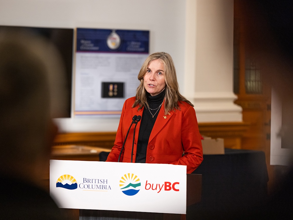 A mid-40s, light-skinned woman with blond hair stands at a lectern. She is wearing a red jacket.
