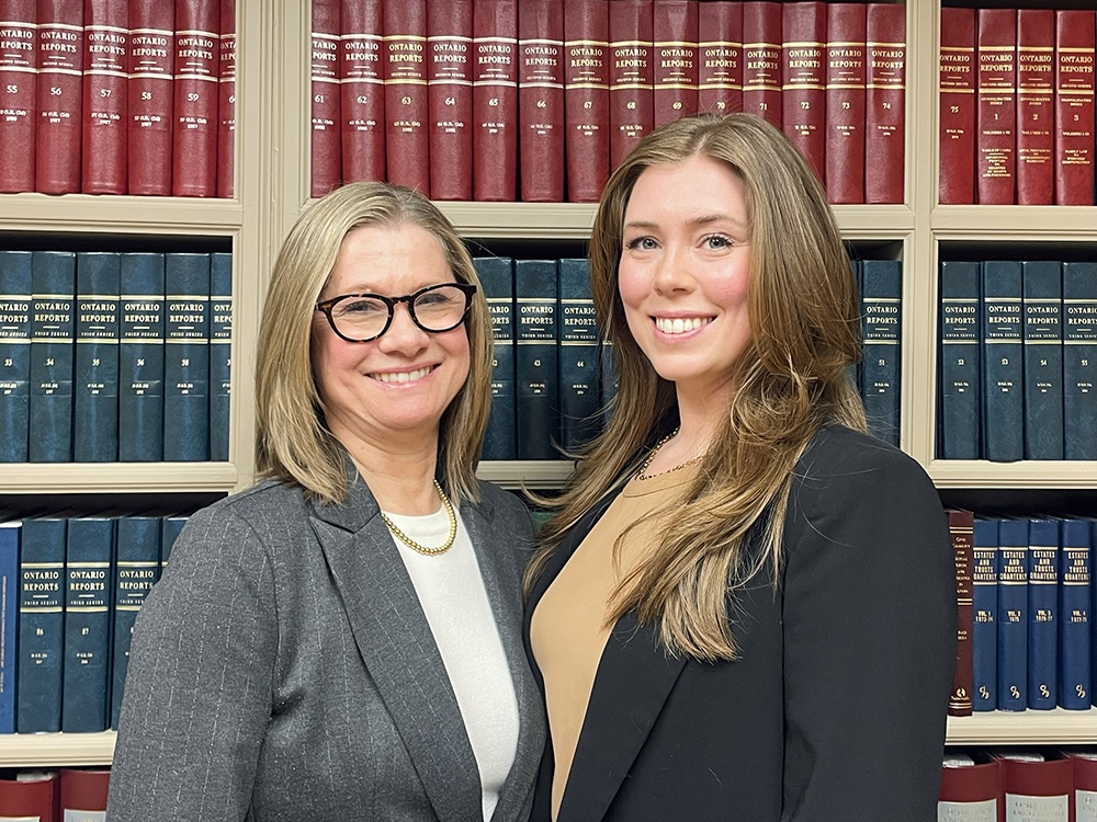 Two women wearing business attire pose in front of a bookshelf. The woman on the left has shoulder-length blond hair and glasses. The woman on the right is taller and has long, light brown hair.