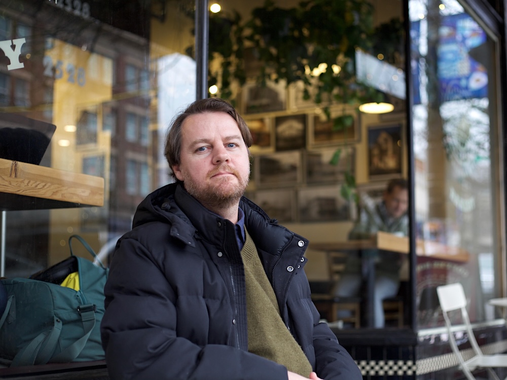 A middle-aged man with light skin and light brown hair, a brown beard and moustache sits at a table outside a caf&eacute;. He wears a puffy black coat over a green sweater.