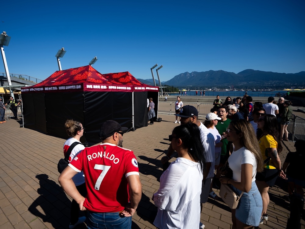 People line up along a boardwalk in Vancouver on a sunny day. In the foreground, a man standing with his back to the camera wears a red &lsquo;Ronaldo&rsquo; jersey. The North Shore mountains can be seen in the background.