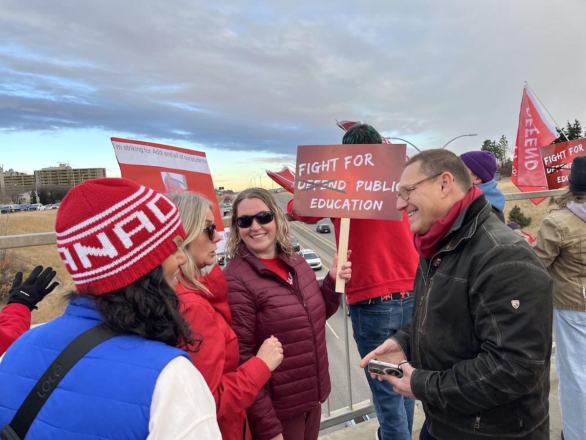 A middle-aged man with light skin and short brown hair speaks with a group of women holding signs with slogans like &lsquo;Fight for public education.&rsquo;