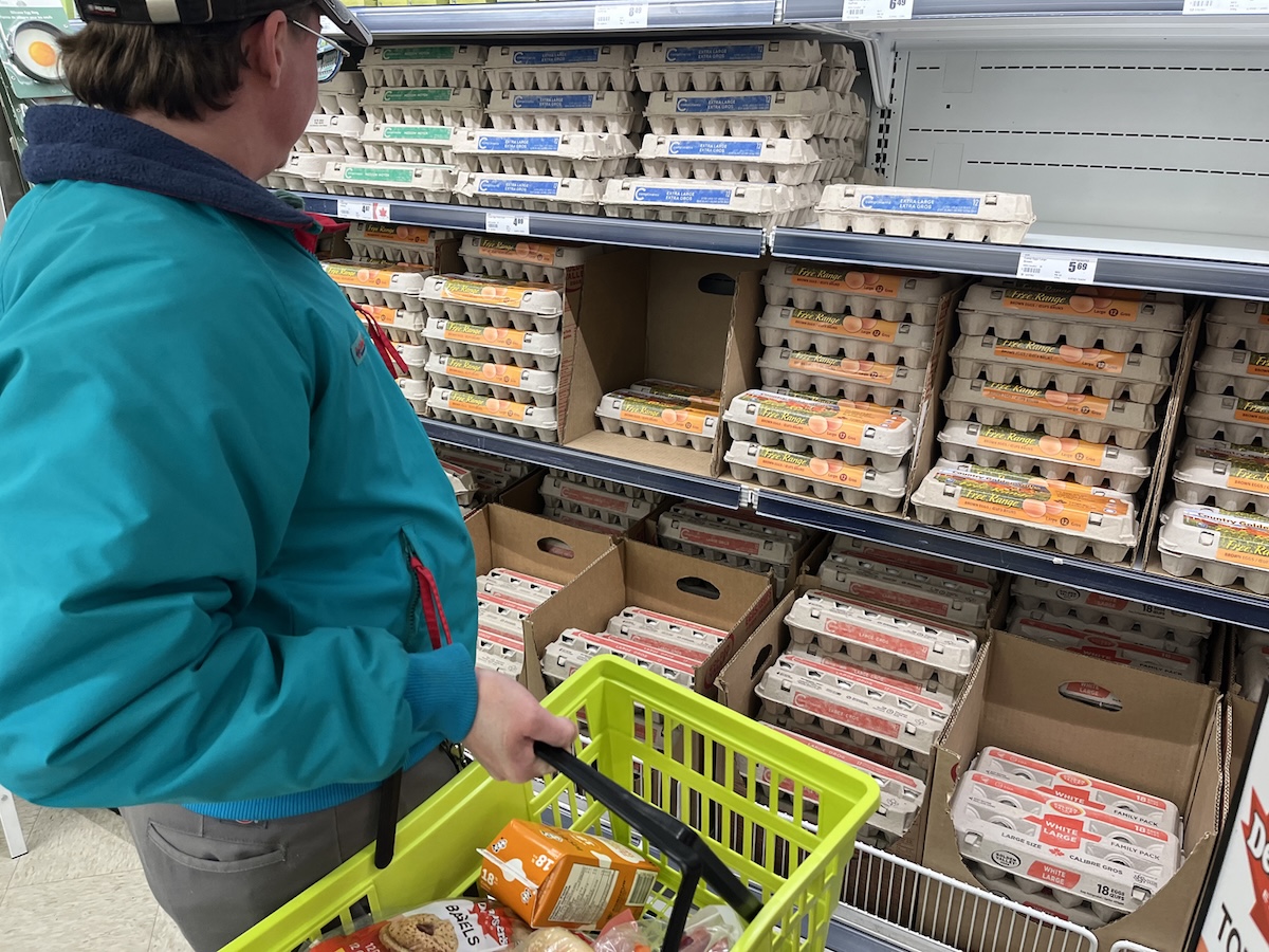A man with light skin tone wearing a ball cap stands in front of the eggs section in the grocery store.