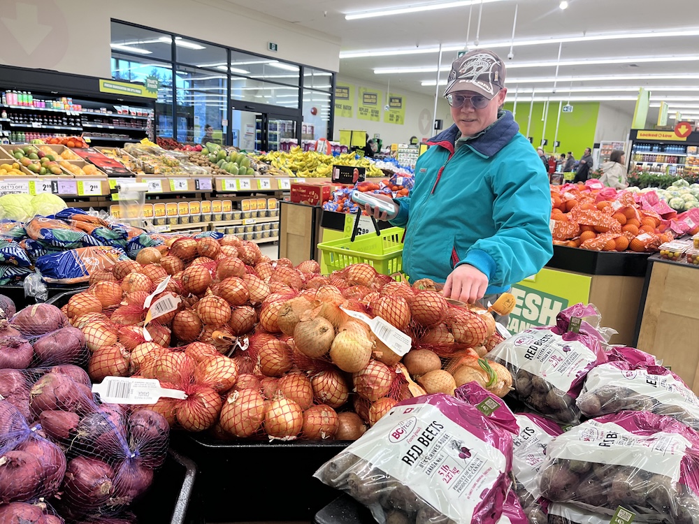 A man with light skin tone wearing a ball cap and turquoise jacket checks out bagged onions in a grocery store.
