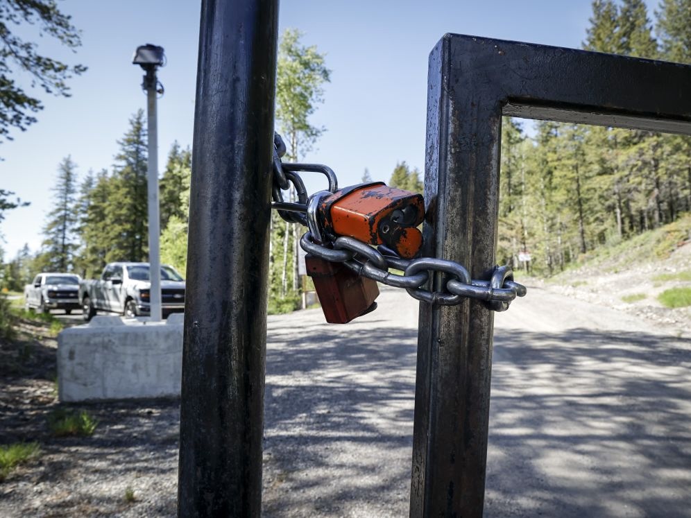 A lock and chain are wrapped around a metal gate in the foreground, with a dirt road and parked security guard vehicles behind it.