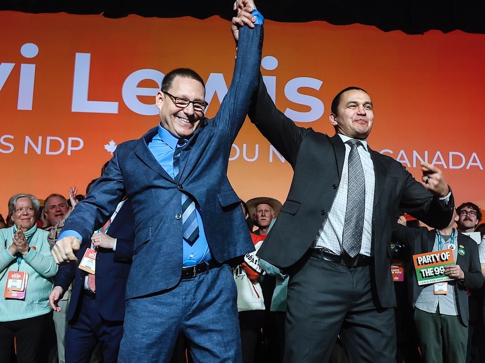 A middle-aged man in a blue suit has his arm hoisted into the air in a victorious gesture by a man in a grey suit as people clap and smile. In the background orange NDP signs can be seen.