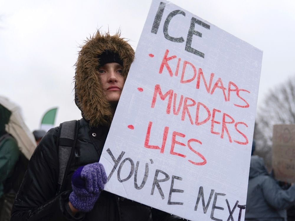 A person with their winter jacket hood up holds a sign that says, &lsquo;ICE kidnaps, murders, lies. You&rsquo;re next.&rsquo;