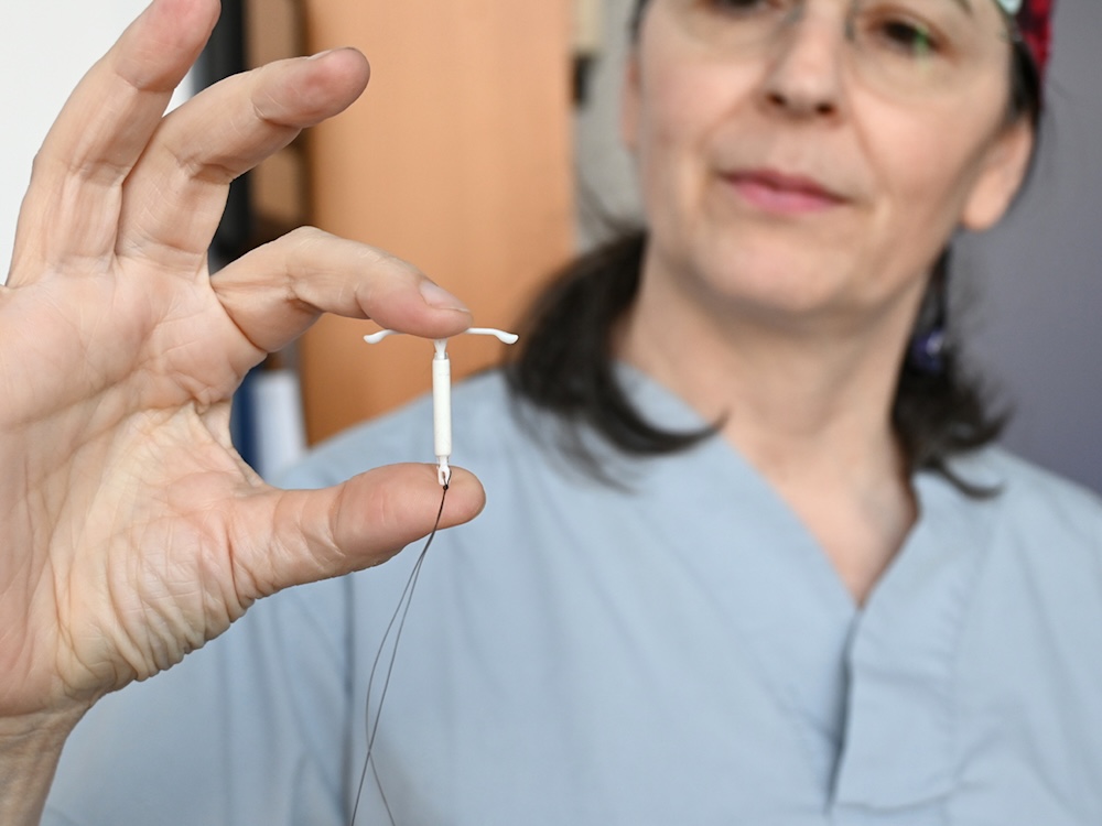 In the foreground, a hand holding a hormonal IUD. Farther back, Dr. Ren&eacute;e Hall, a woman with shoulder-length dark hair and light skin tone who is wearing blue scrubs.