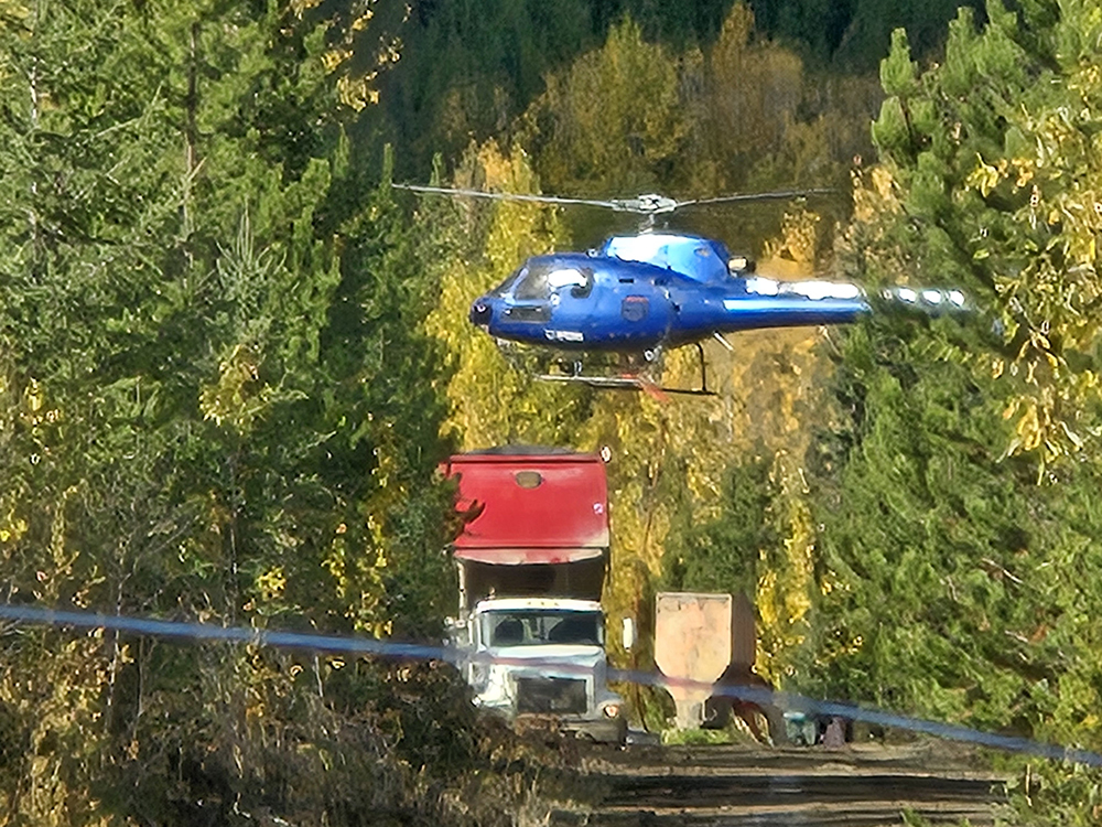 A helicopter hovers near the ground. Below there is a large truck and other equipment.