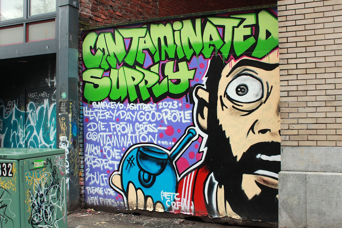 A graffiti mural reads, &lsquo;Contaminated Supply.&rsquo; A person with a beard holds a glass pipe.
