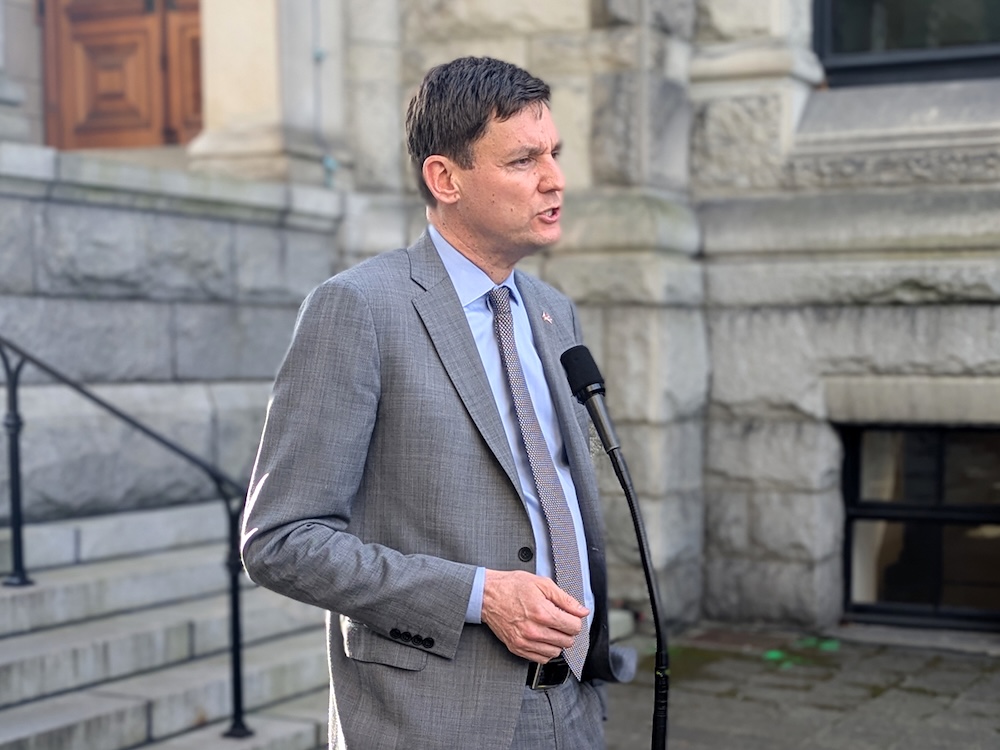 David Eby, a tall man with light skin tone, stands at a microphone in front of the legislature.