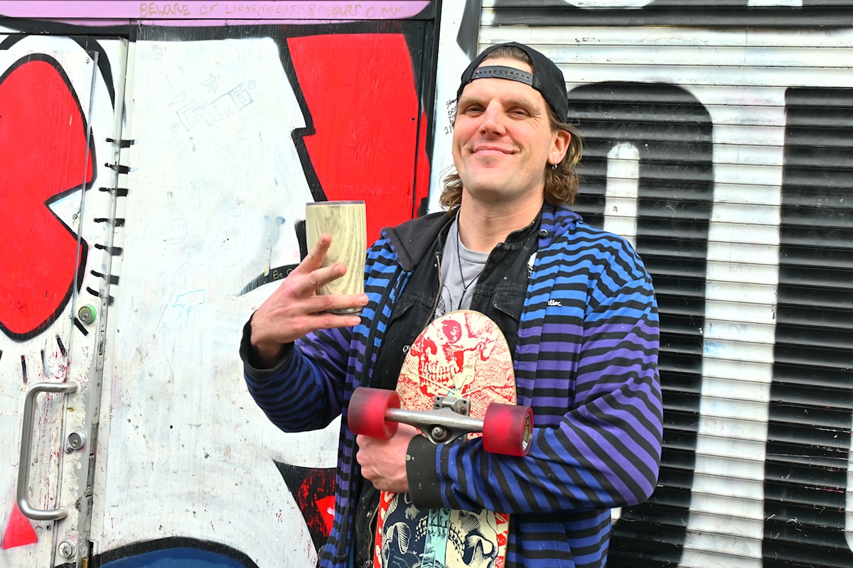 A man with a backwards baseball cap and a blue and purple striped sweatshirt grins at the camera. He&rsquo;s holding a skateboard and a coffee cup and standing in front of bold graffiti.
