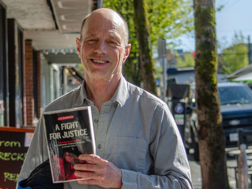 A light-skinned man in a grey button-front shirt smiles, holding a red book entitled &ldquo;A Fight For Justice&rdquo; while standing next to an orange brick wall.