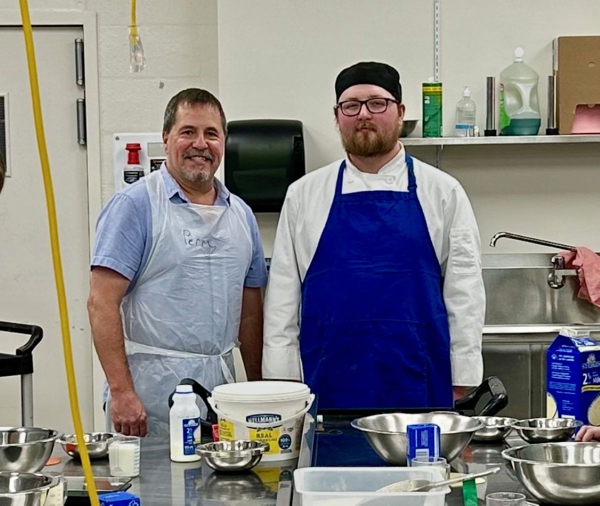 A middle-aged man wearing an apron poses beside a young man in a beard, glasses and apron. Bowls and baking ingredients can be seen on the counter in front of the two men.