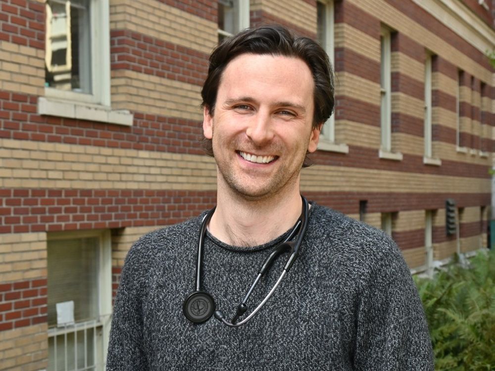 A man with light skin tone and dark hair poses for a photo outside. He is wearing a speckled wool sweater and a stethoscope.