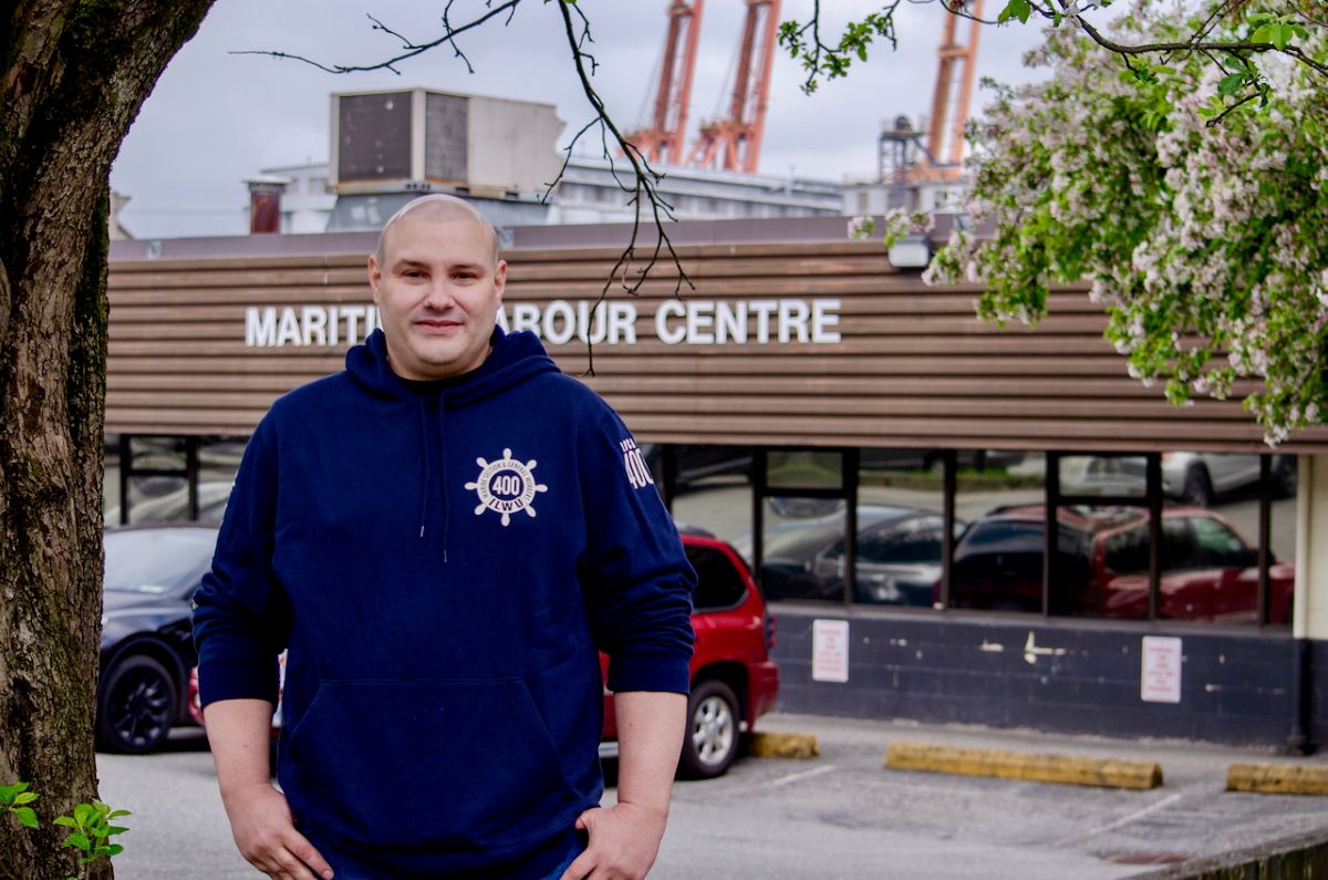 A man wearing a dark blue sweatshirt stands in front of a building with a sign that reads &ldquo;Maritime Labour Centre.&rdquo;