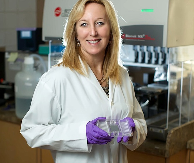 A woman with light skin, blond hair, a white lab coat and purple gloves holds up a slide with organized rows of dots. She is standing in a lab in front of machinery. 